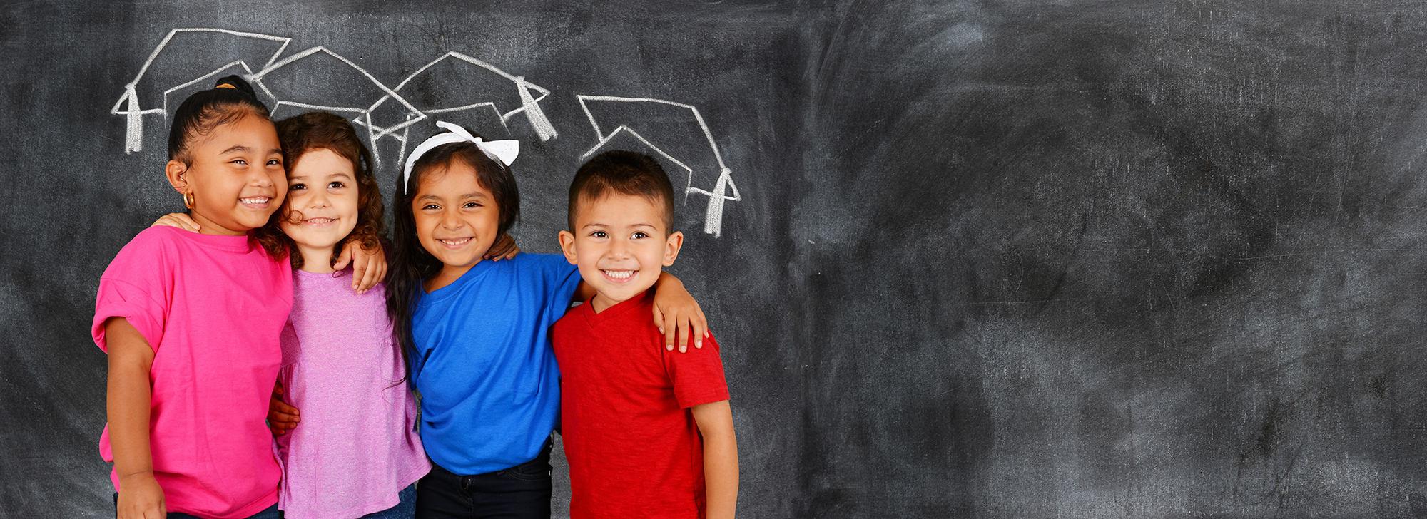 four children standing against a chalkboard with graduation caps drawn above their heads